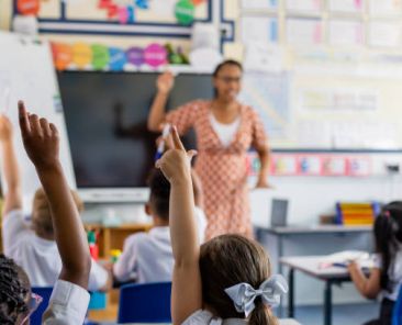 Primary school students sitting in a classroom being taught by a teacher in the North East of England. The students are answering questions with their arms in the air, the focus being on their arms.